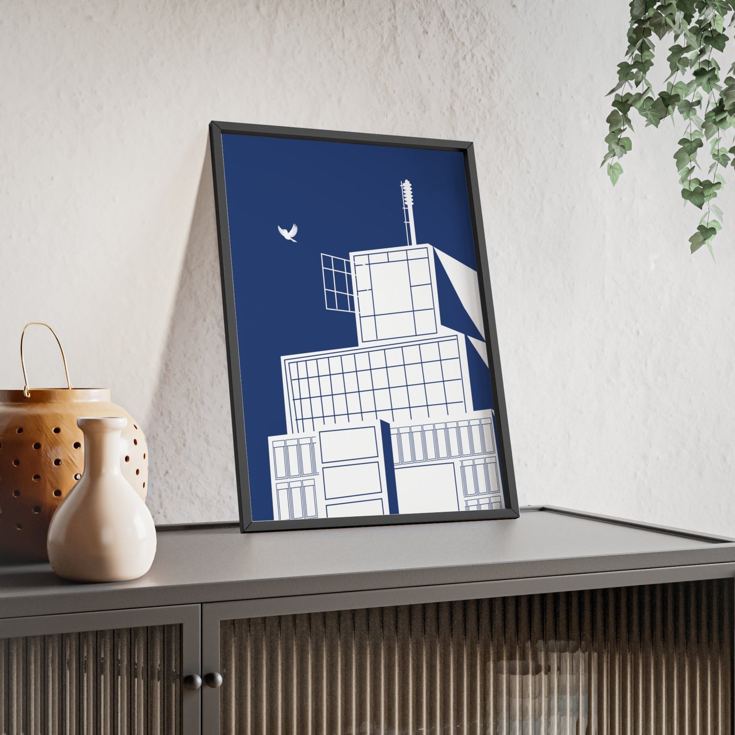 Framed geometric building art on a blue background, placed on a cabinet with vases and leafy decor.