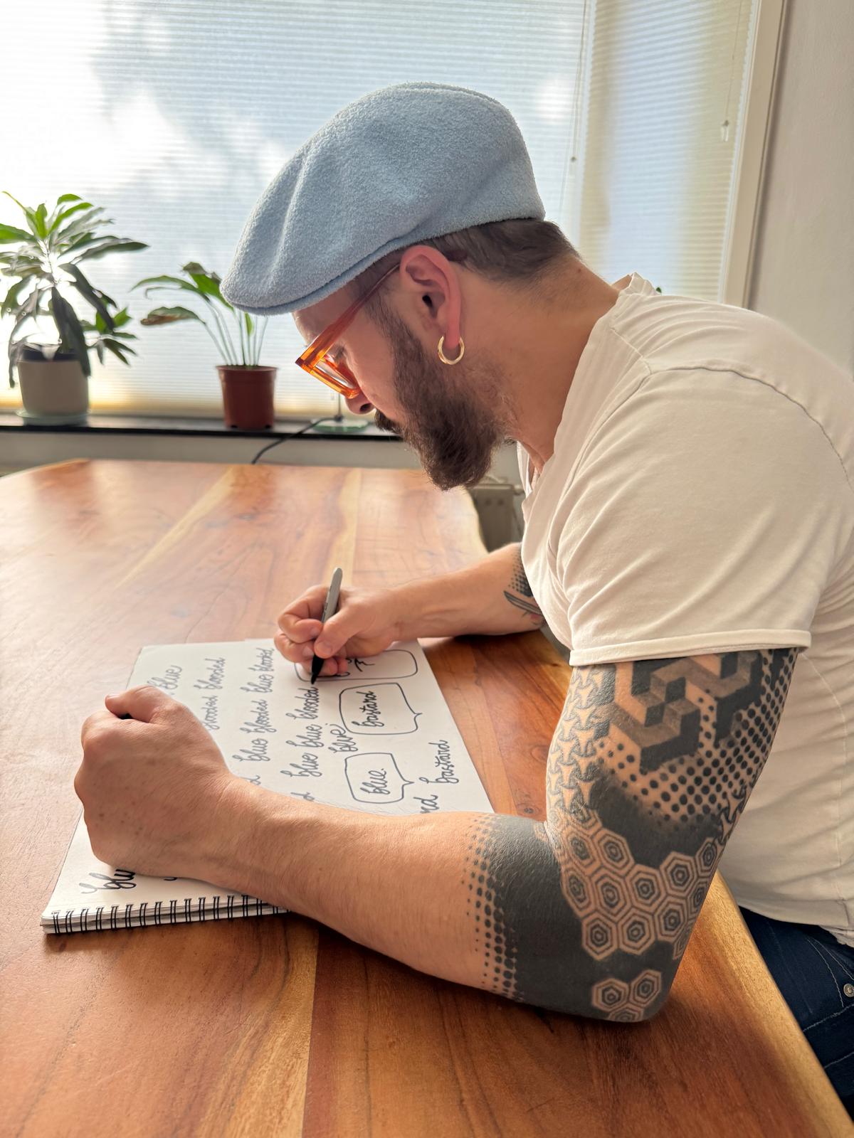 Man with a blue cap and tattoos, writing on paper at a wooden table with potted plants nearby.